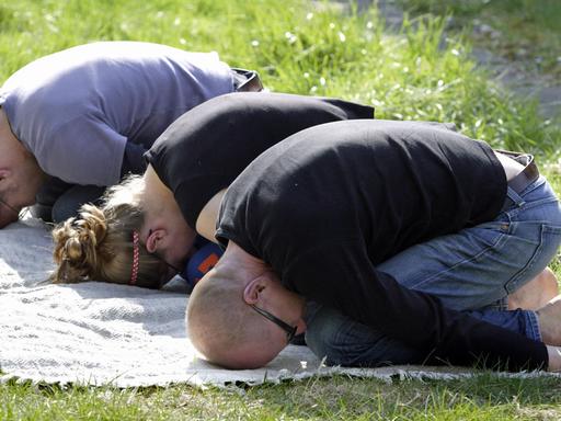 Drei junge Menschen bei der Gymnastik im Park: Sie sitzen zusammengekauert auf den Knien, die Arme nach hinten auf dem Boden gestreckt und die Stirn gegen den Untergrund gedrückt.