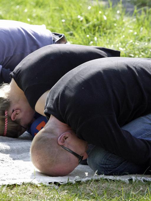 Drei junge Menschen bei der Gymnastik im Park: Sie sitzen zusammengekauert auf den Knien, die Arme nach hinten auf dem Boden gestreckt und die Stirn gegen den Untergrund gedrückt.
