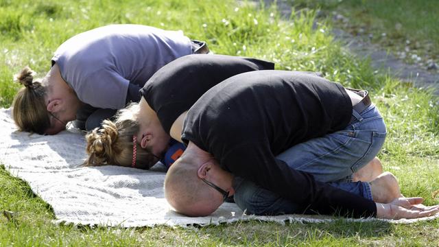 Drei junge Menschen bei der Gymnastik im Park: Sie sitzen zusammengekauert auf den Knien, die Arme nach hinten auf dem Boden gestreckt und die Stirn gegen den Untergrund gedrückt.