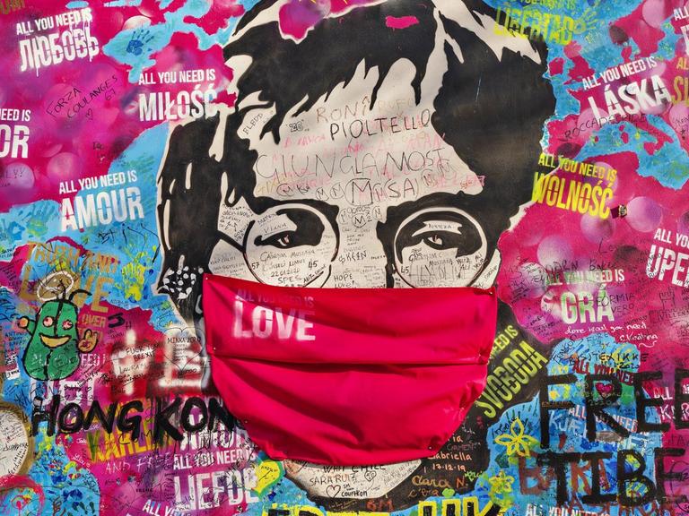 John Lennon-Graffito mit Atemschutz auf einer Weltkarte mit "All you need is love"-Sprüchen