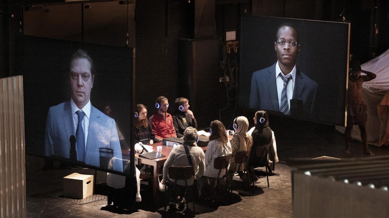 Szenenfoto: Acht Menschen sitzen mit Kopfhörern an einem Tisch. Rechts und links von ihnen sind zwei Projektionen zu sehen: ein weißer Mann und ein dunkelhäutiger Mann.