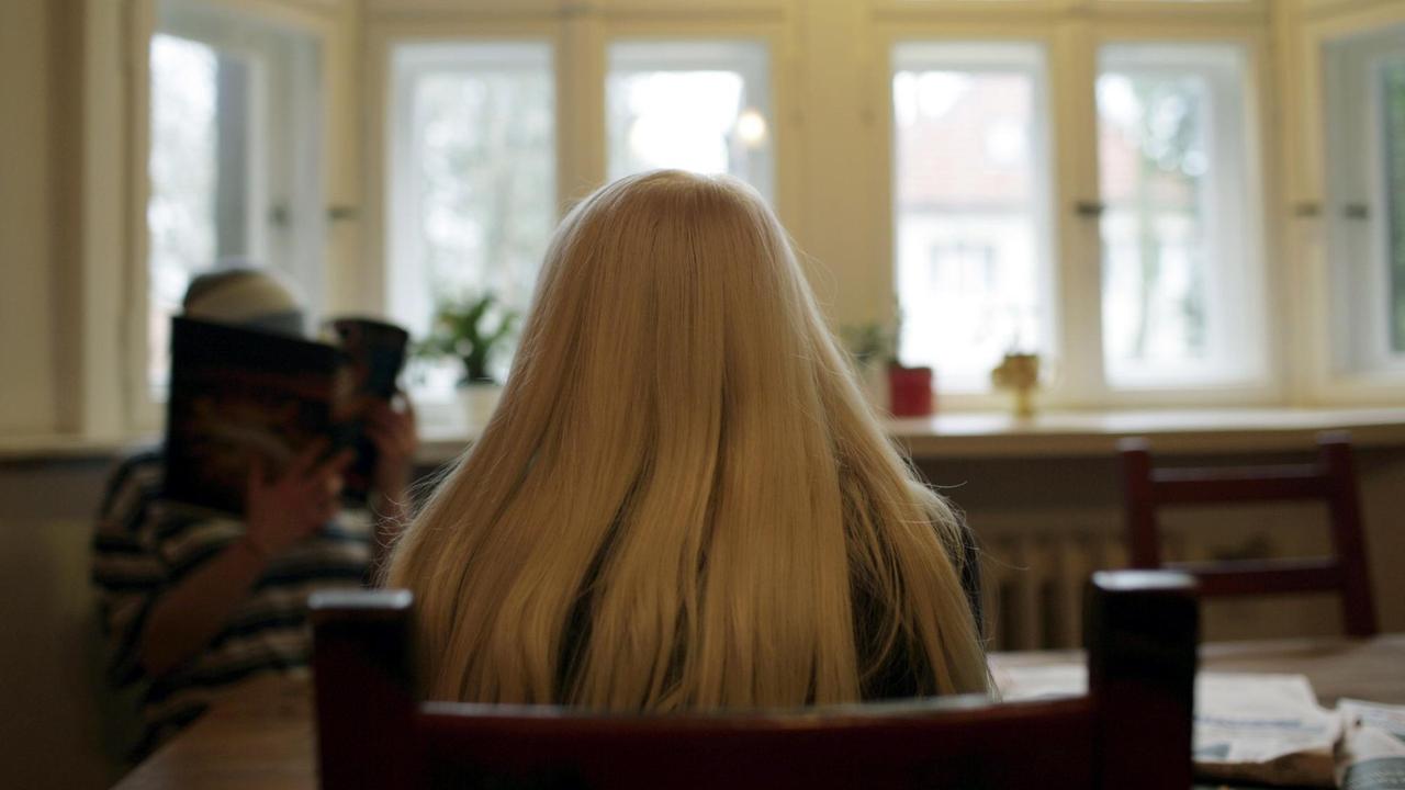 Eine Frau mit langen blonden Haaren sitzt in einem großen Raum mit hohen Fenstern. Eine Frau mit langen blonden Haaren sitzt in einem großen Raum mit hohen Fenstern.