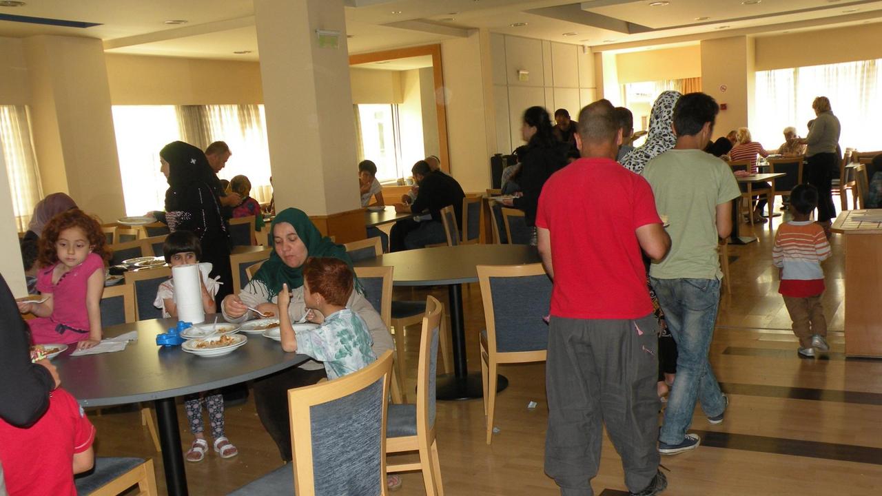 Farbfoto eines Innenraums, in dem Familien aus arabischen Ländern beim Essen sitzen. Flüchtlinge in einer Flüchtlingsunterkunft