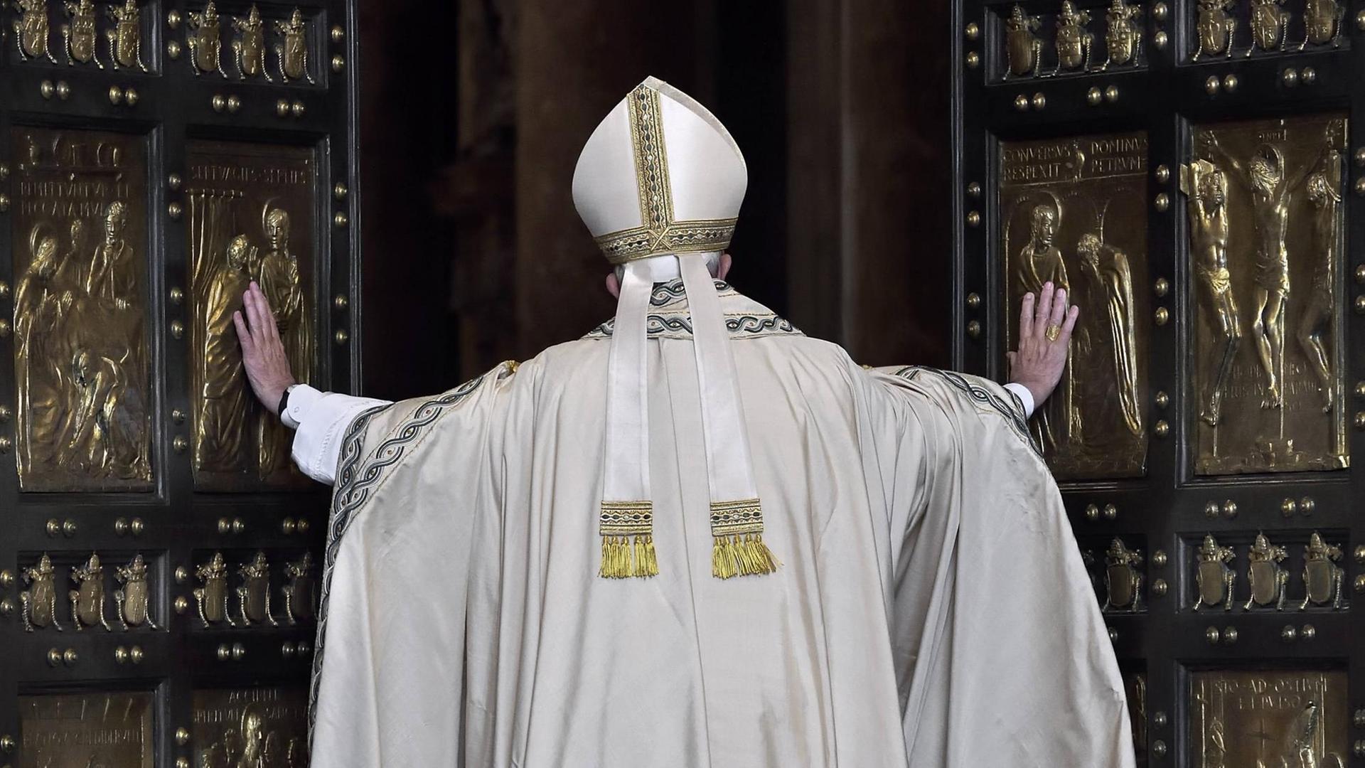 Papst Franziskus von Hinten, wie er die großen Flügel der "Heiligen Pforte" öffnet. Das Portal besteht aus zwei großen Kassettentüren mit schwarzen Rahmen und goldenen Kassetten mit christlichen Motiven. Papst Franziskus von Hinten, wie er die großen Flügel der "Heiligen Pforte" öffnet. Das Portal besteht aus zwei großen Kassettentüren mit schwarzen Rahmen und goldenen Kassetten mit christlichen Motiven.