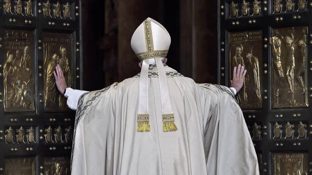 Papst Franziskus von Hinten, wie er die großen Flügel der "Heiligen Pforte" öffnet. Das Portal besteht aus zwei großen Kassettentüren mit schwarzen Rahmen und goldenen Kassetten mit christlichen Motiven.