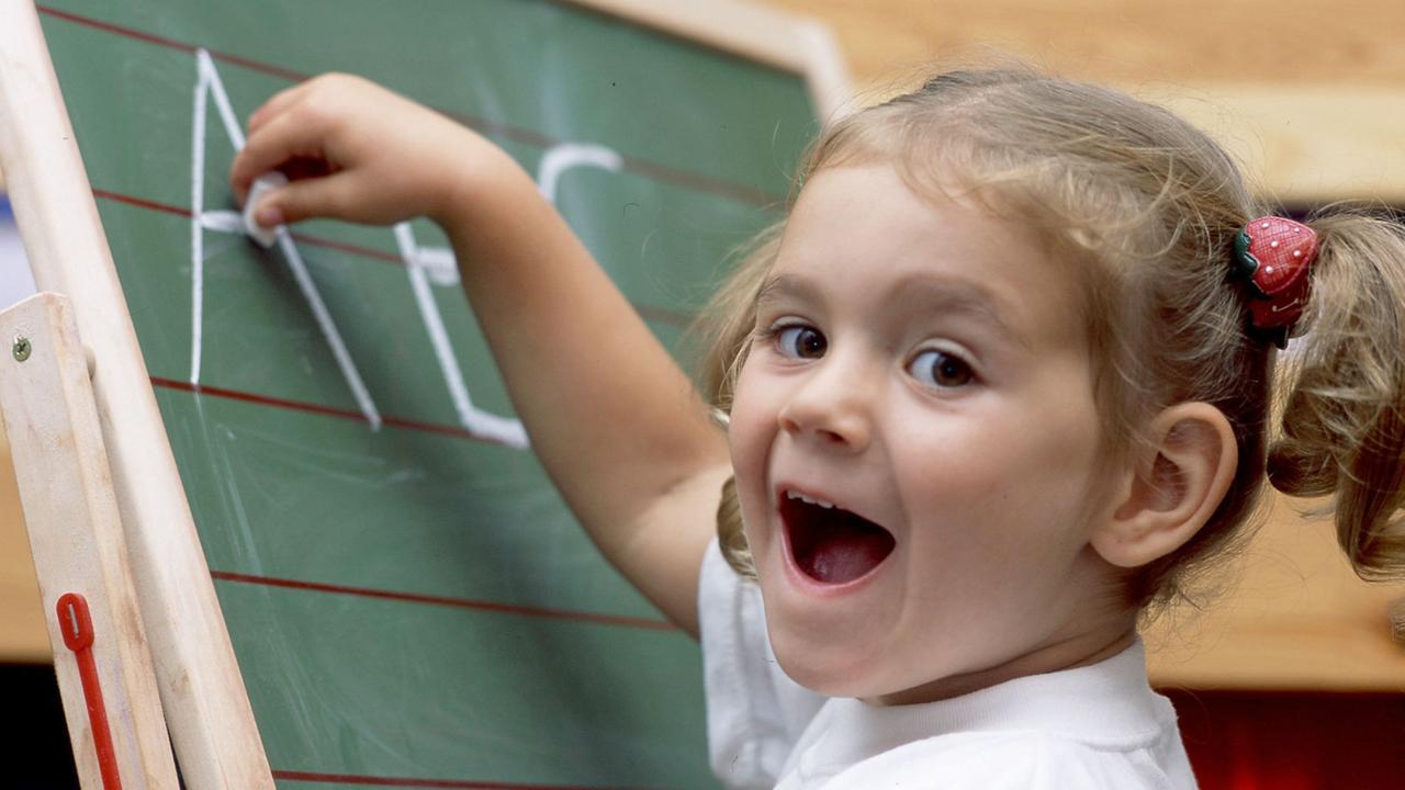 Mit Eifer schreibt ein fünfjähriges Mädchen im weißen T-Shirt und mit Zöpfen bei dem gestellten Symbolbild die Buchstaben A B und C mit Kreide auf eine Schultafel. Mit Eifer schreibt ein fünfjähriges Mädchen im weißen T-Shirt und mit Zöpfen bei dem gestellten Symbolbild die Buchstaben A B und C mit Kreide auf eine Schultafel.
