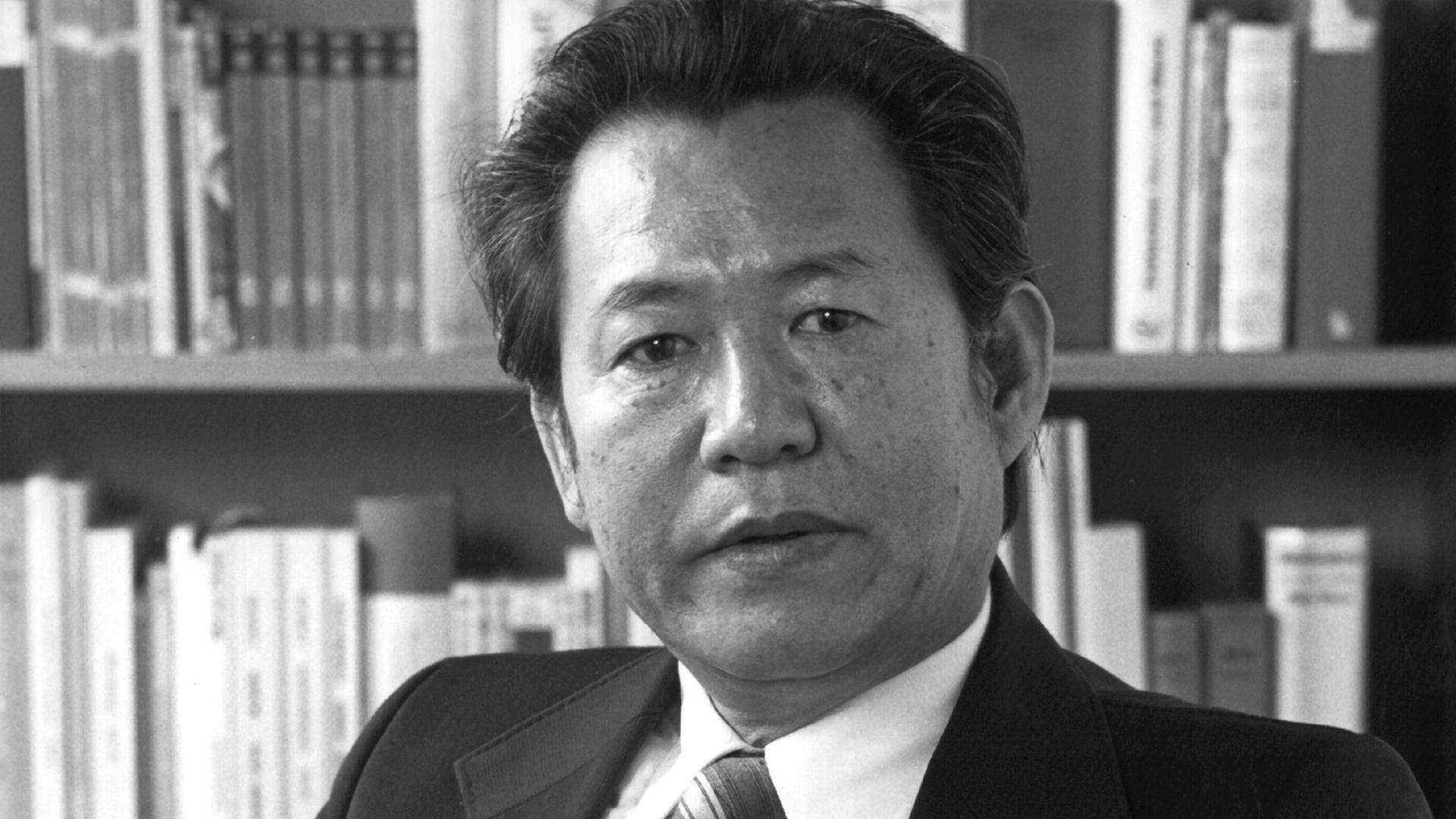 Ein mittelalter Mann mit asiatischem Aussehen blickt auf einer Schwarzweißporträtaufnahme in die Kamera, hinter ihm stehen Bücher in einem Regal. Ein mittelalter Mann mit asiatischem Aussehen blickt auf einer Schwarzweißporträtaufnahme in die Kamera, hinter ihm stehen Bücher in einem Regal.