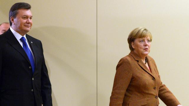 Viktor Janukowitsch und Angela Merkel gehen hintereinander vor einer hellen Wand entlang.
