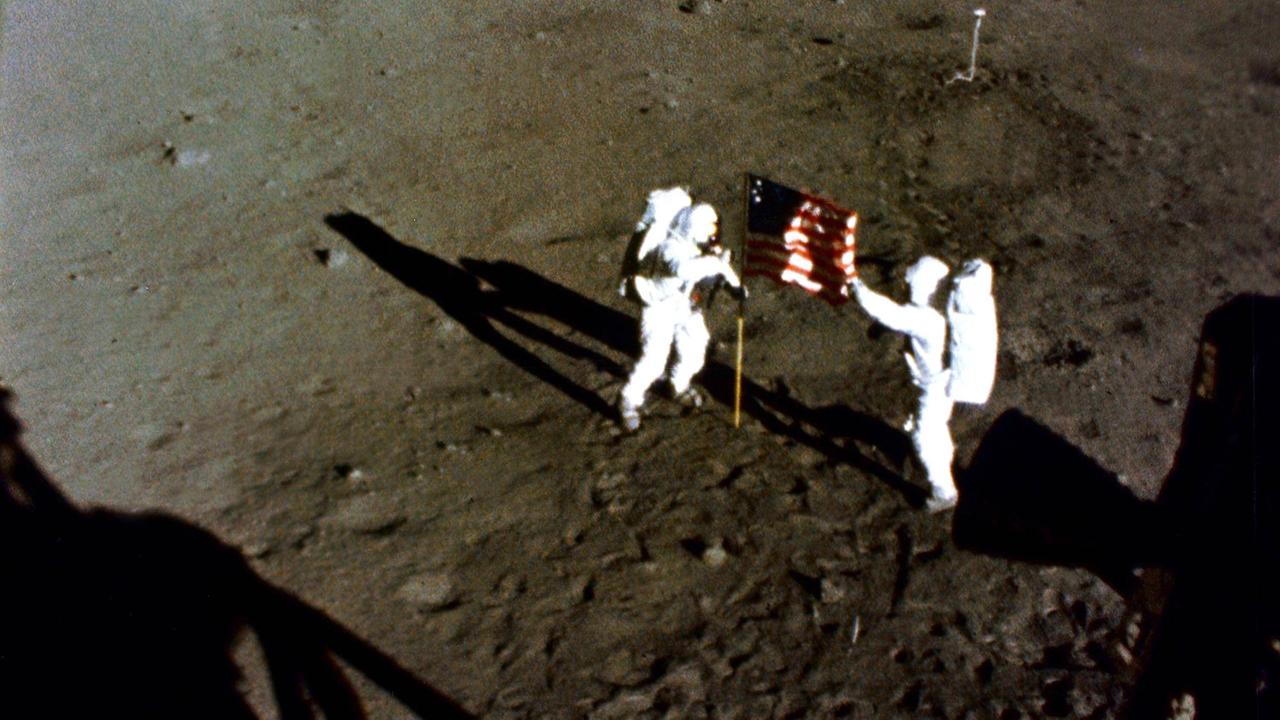 Buzz Aldrin und Neil Armstrong stehen in Raumanzügen auf der Mondoberfläche, zwischen ihnen die Flagge der USA. Buzz Aldrin und Neil Armstrong stehen in Raumanzügen auf der Mondoberfläche, zwischen ihnen die Flagge der USA.