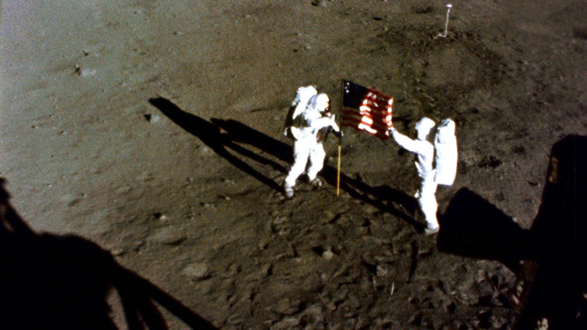 Buzz Aldrin und Neil Armstrong stehen in Raumanzügen auf der Mondoberfläche, zwischen ihnen die Flagge der USA. Buzz Aldrin und Neil Armstrong stehen in Raumanzügen auf der Mondoberfläche, zwischen ihnen die Flagge der USA.
