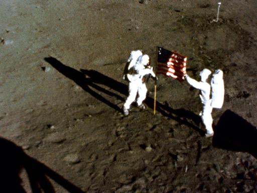 Buzz Aldrin und Neil Armstrong stehen in Raumanzügen auf der Mondoberfläche, zwischen ihnen die Flagge der USA.
