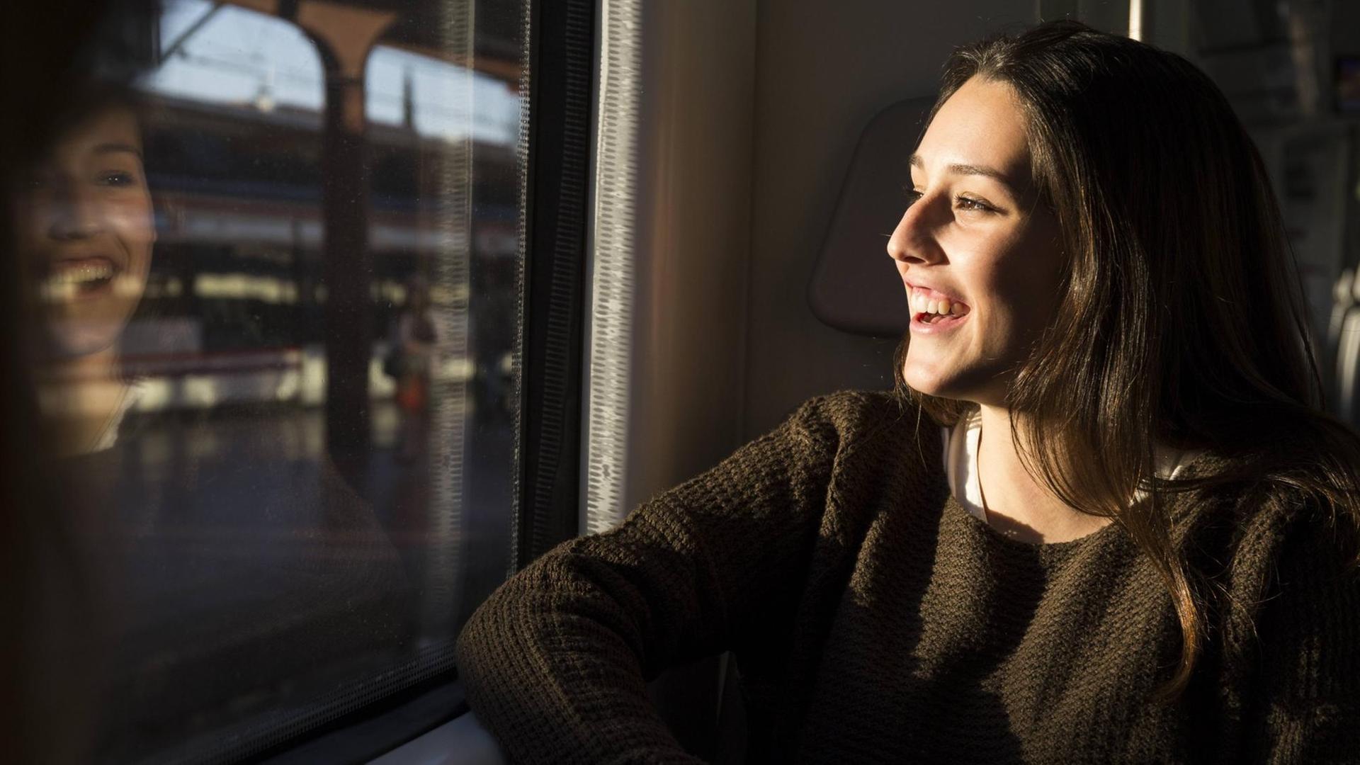 Eine fröhliche junge Frau sitzt in einem Zug und schaut aus dem Fenster auf den vorbeiziehenden Bahnhof. In der Scheibe spiegelt sich ihr lachendes Gesicht. Eine fröhliche junge Frau sitzt in einem Zug und schaut aus dem Fenster auf den vorbeiziehenden Bahnhof. In der Scheibe spiegelt sich ihr lachendes Gesicht.