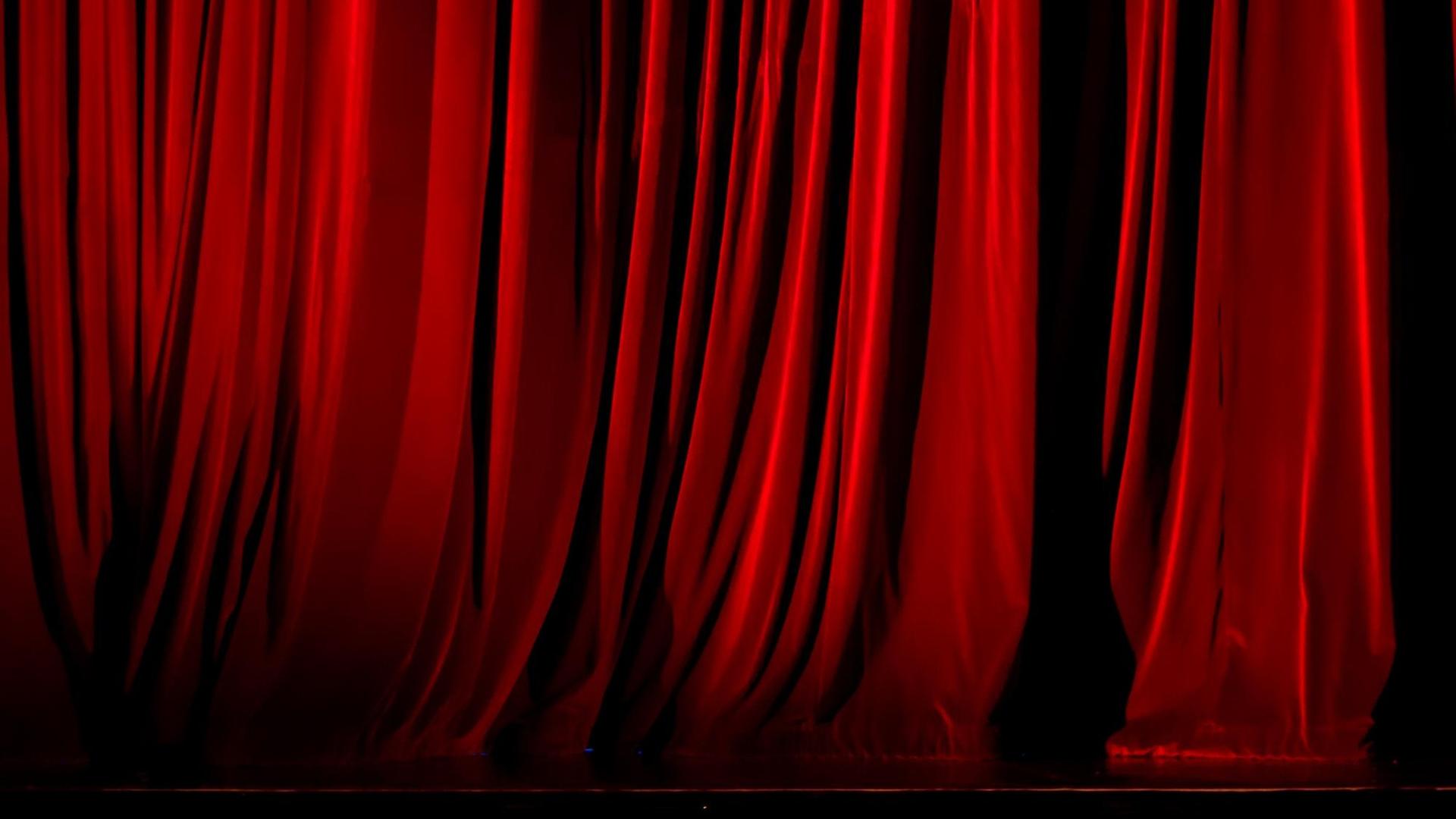 Zu sehen ist ein beleuchteter roter Vorhang auf einer Theater- oder Opernbühne. Zu sehen ist ein beleuchteter roter Vorhang auf einer Theater- oder Opernbühne.