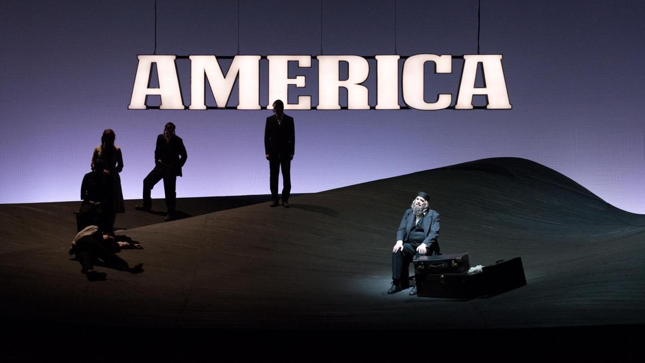 Auf der dunkel ausgeleuchteten Bühne prangt der beleuchtete Schriftzug "America" hinter dem Darsteller des Tora-Lehrers Mendel Singer . Auf der dunkel ausgeleuchteten Bühne prangt der beleuchtete Schriftzug "America" hinter dem Darsteller des Tora-Lehrers Mendel Singer .