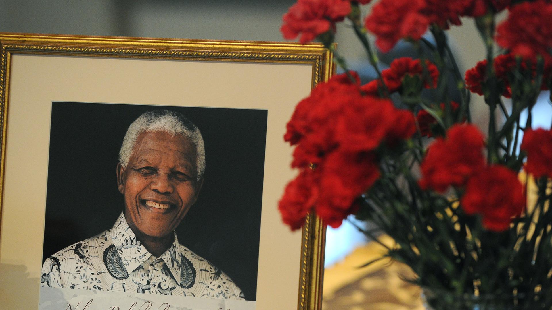 Neben einem Portraitbild Nelson Mandelas steht eine Vase roter Nelken.