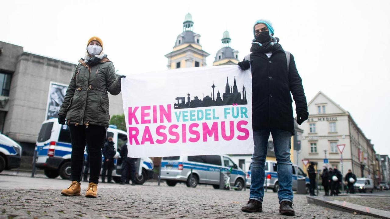 Zwei Demonstrierende mit Mund-Nasen-Schutz halten ein Plakat mit der Aufschrift „Kein Veedel für Rassismus“ hoch. Zwei Demonstrierende mit Mund-Nasen-Schutz halten ein Plakat mit der Aufschrift „Kein Veedel für Rassismus“ hoch.