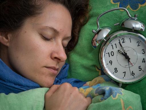 Eine Frau schläft im Bett, neben ihr liegt ein Wecker.