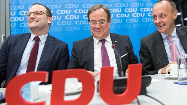 Jens Spahn, Armin Laschet und Friedrich Merz sitzen an einem Tisch, im Vordergrund sieht man groß die roten Buchstaben "CDU"