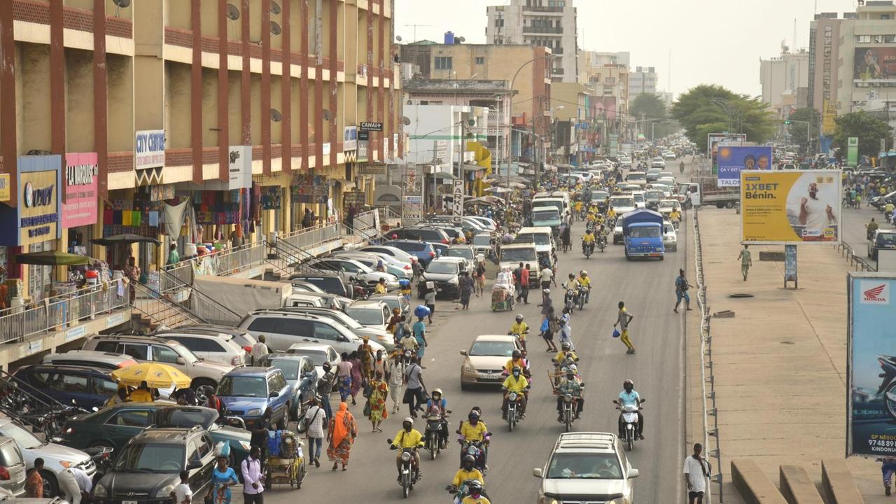 Cotonou ist mit seinen geschätzt 800.000 Einwohnern das Wirtschaftszentrum von Benin. Über eine große Straße rasen die typischen Zémidjans, Moped-Taxen, durch den dichten Verkehr.. Cotonou ist mit seinen geschätzt 800.000 Einwohnern das Wirtschaftszentrum von Benin. Über eine große Straße rasen die typischen Zémidjans, Moped-Taxen, durch den dichten Verkehr..