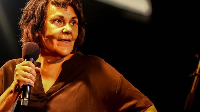 Die Sängerin Christiane Rösinger steht auf einer Bühne. In der rechten Hand hält sie ein Mikrofon. Die Sängerin Christiane Rösinger steht auf einer Bühne. In der rechten Hand hält sie ein Mikrofon.