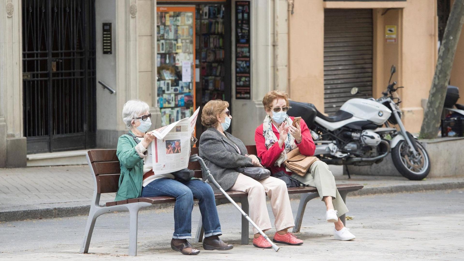 Drei ältere Frauen sitzen in mit Coronamaske in Barcelona auf einer Bank. Eine Frau liest Zeitung, die beiden anderen unterhalten sich. Drei ältere Frauen sitzen in mit Coronamaske in Barcelona auf einer Bank. Eine Frau liest Zeitung, die beiden anderen unterhalten sich.