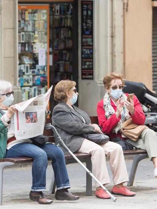 Drei ältere Frauen sitzen in mit Coronamaske in Barcelona auf einer Bank. Eine Frau liest Zeitung, die beiden anderen unterhalten sich.