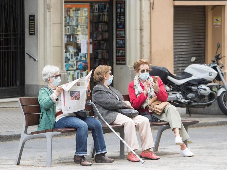 Drei ältere Frauen sitzen in mit Coronamaske in Barcelona auf einer Bank. Eine Frau liest Zeitung, die beiden anderen unterhalten sich.