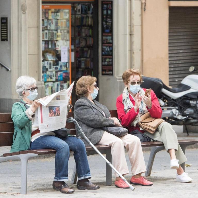 Drei ältere Frauen sitzen in mit Coronamaske in Barcelona auf einer Bank. Eine Frau liest Zeitung, die beiden anderen unterhalten sich. 