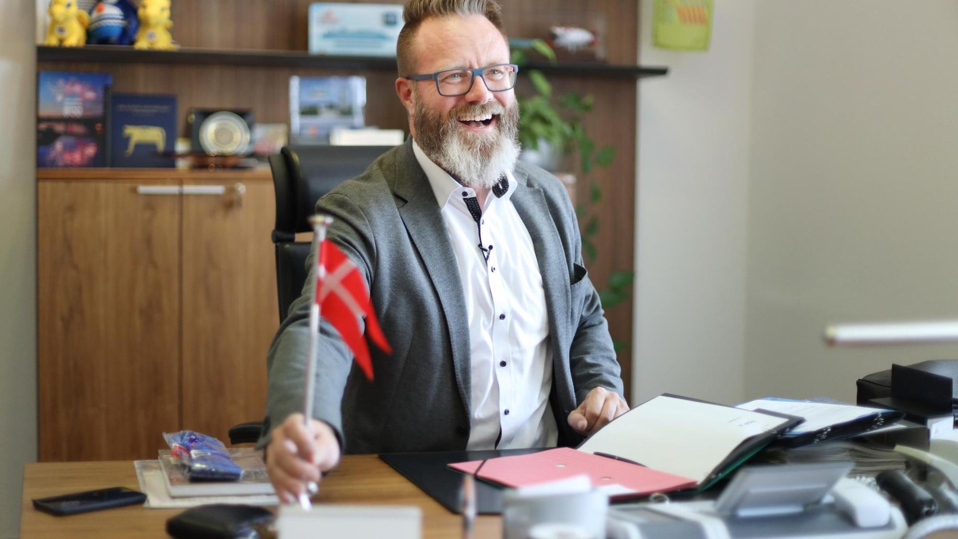 Rostocks neuer Oberbürgermeister Claus Ruhe Madsen sitzt am Schreibtisch seines Büro. Er lacht ind hält in seiner rechten Hand die dänische Flagge. Rostocks neuer Oberbürgermeister Claus Ruhe Madsen sitzt am Schreibtisch seines Büro. Er lacht ind hält in seiner rechten Hand die dänische Flagge.