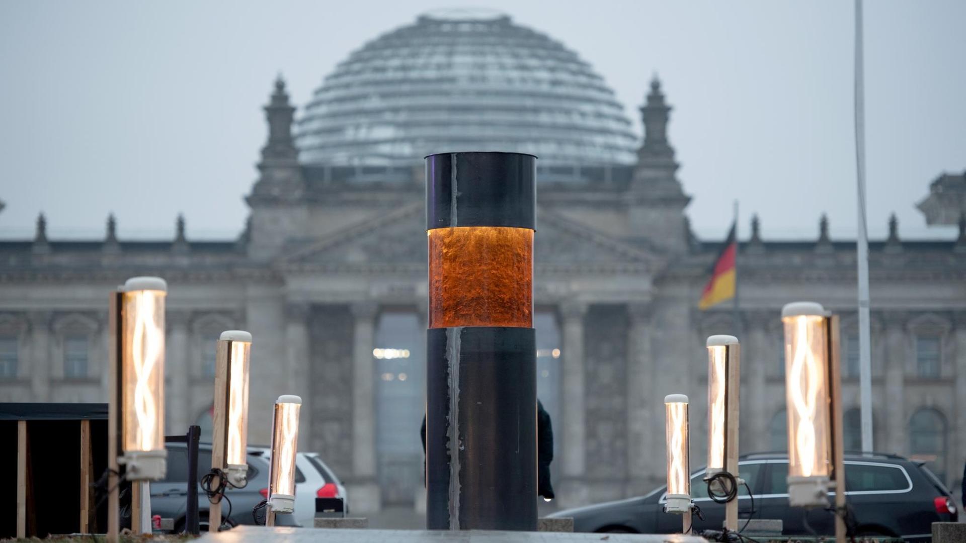 Die Säule aus Metall hat im oberen Drittel eine rötliche Glasscheibe, durch die man die Asche sieht. Im Hintergrund unscharf der Reichstag.