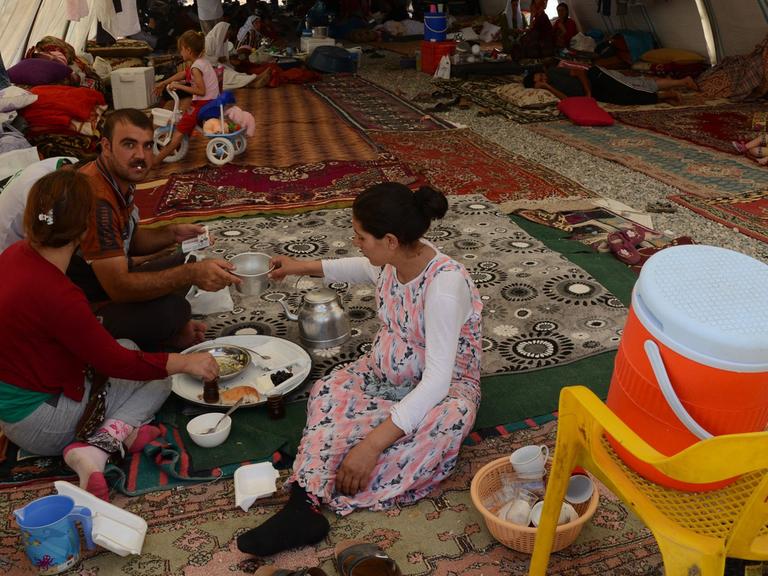 Eine yezidische Familie hat Schutz gefunden in einem Flüchtlingscamp in der Nähe der türkisch-irakischen Grenze. Erwachsene und Kinder sitzen auf ausgebreiteten Teppichen in einem Zelttunnel.