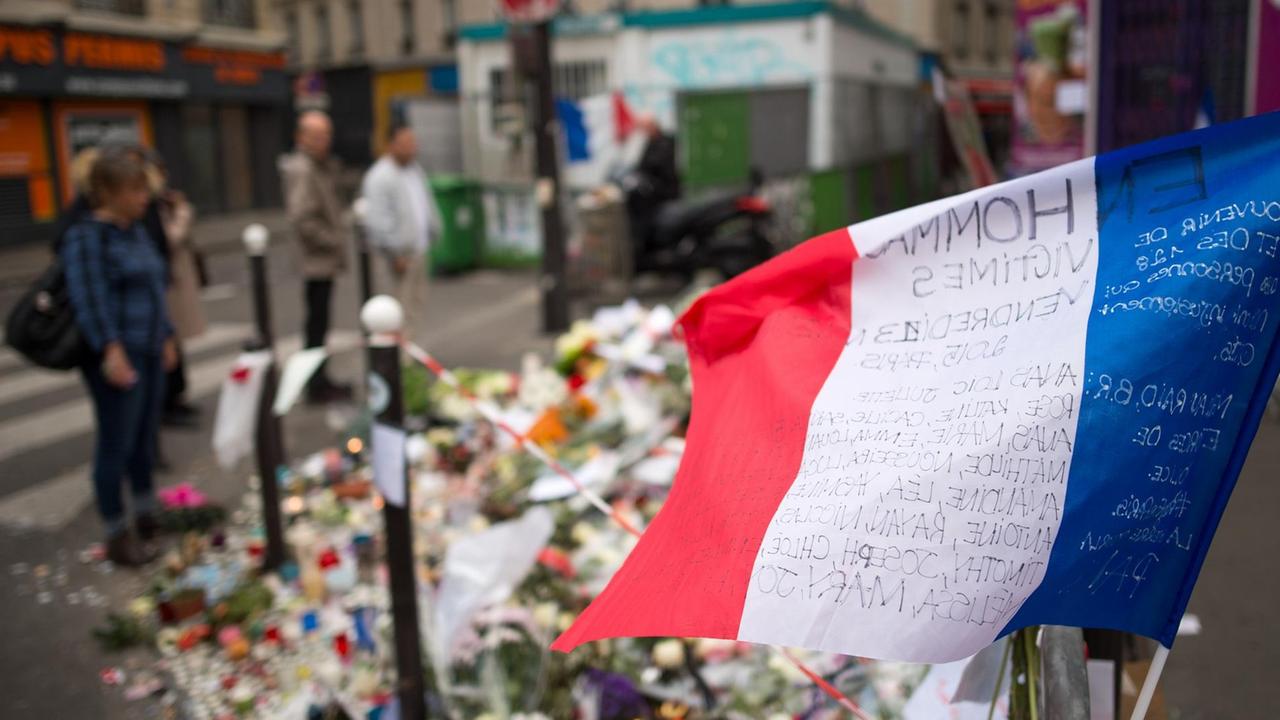 Eine Frankreich-Fahne in blau-weiß-rot in der Nähe des Konzertsaals Bataclan in Paris, im Hintergrund liegen Blumen auf dem Bürgersteig.