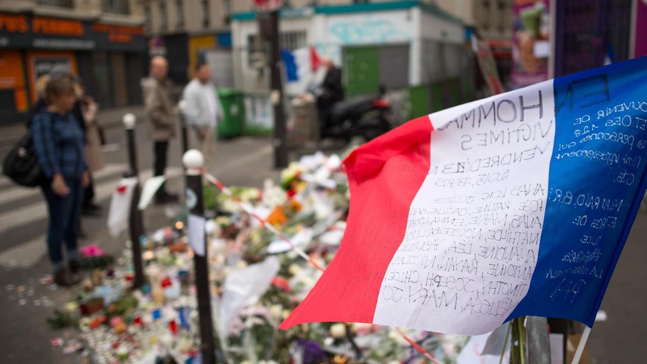 Eine Frankreich-Fahne in blau-weiß-rot in der Nähe des Konzertsaals Bataclan in Paris, im Hintergrund liegen Blumen auf dem Bürgersteig. Eine Frankreich-Fahne in blau-weiß-rot in der Nähe des Konzertsaals Bataclan in Paris, im Hintergrund liegen Blumen auf dem Bürgersteig.
