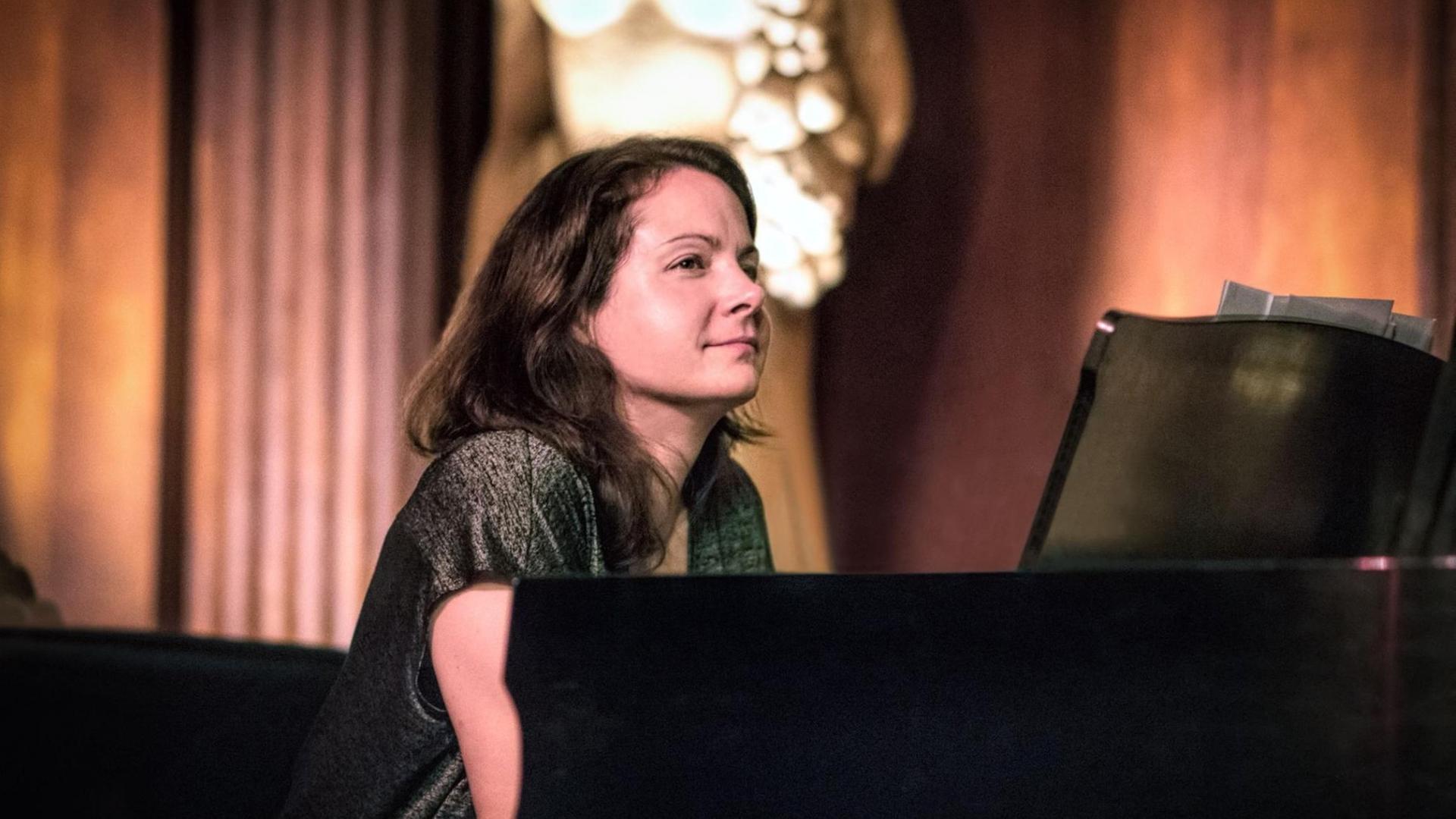 Vor einem schwarzen Piano sitzt eine dunkelhaarige junge Frau. Ihr Blick schweift zur Seite und im Hintergrund ist verschwommen eine Statue zu sehen. Vor einem schwarzen Piano sitzt eine dunkelhaarige junge Frau. Ihr Blick schweift zur Seite und im Hintergrund ist verschwommen eine Statue zu sehen.