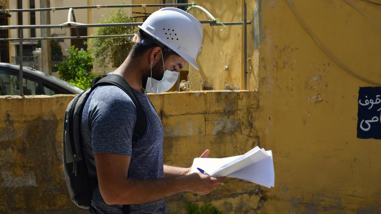 Der 26-jährige Architekt Bahaa Baschnak mit Helm und Maske vor einem senfgelben Haus