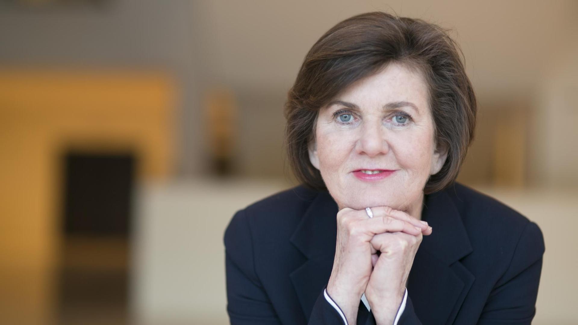 Eine Dame mit kurzen braunen Haaren und blauem Blazer stützt das Kinn auf die gefaltete Faust. Eine Dame mit kurzen braunen Haaren und blauem Blazer stützt das Kinn auf die gefaltete Faust.