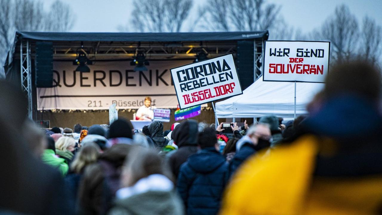 Eine Menschenmenge steht vor einer Bühne. Einige tragen Plakate, etwa mit der Botschaft: "Corona Rebellen Düsseldorf", und "Wir sind der Souverän". Eine Menschenmenge steht vor einer Bühne. Einige tragen Plakate, etwa mit der Botschaft: "Corona Rebellen Düsseldorf", und "Wir sind der Souverän".