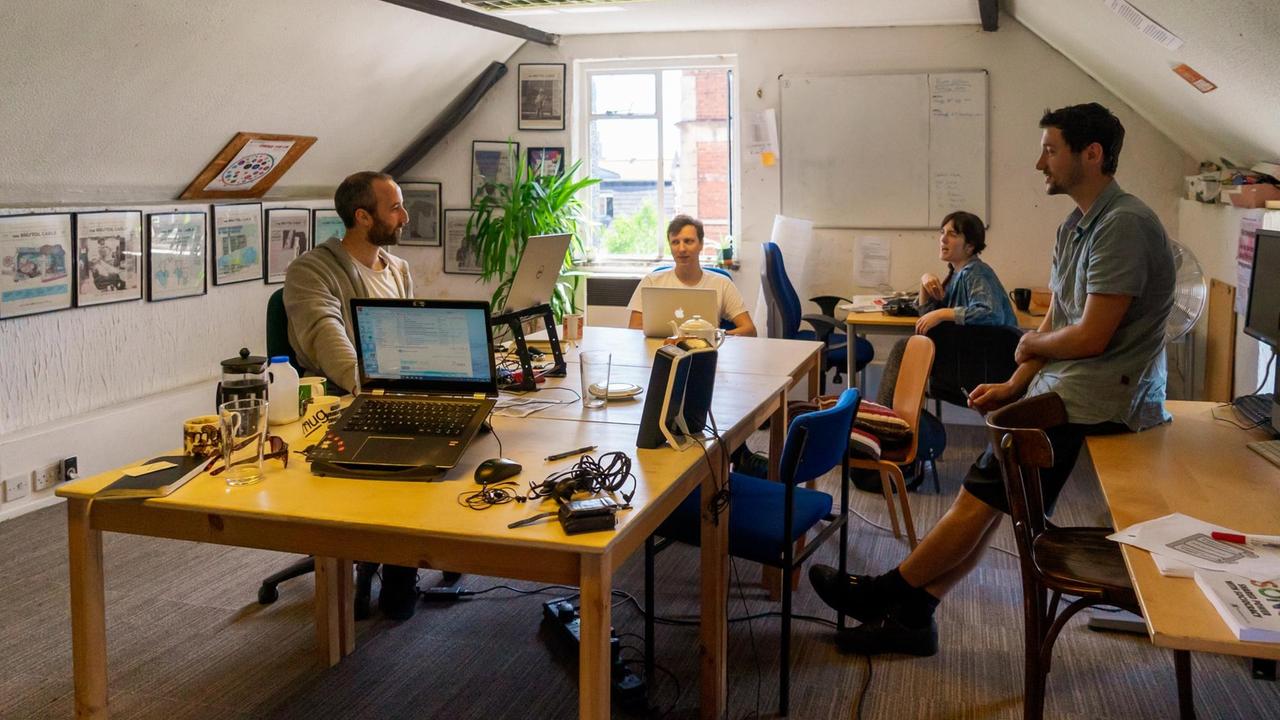 Vier Männer und Frauen sitzen und lehnen an Tischen in der Redaktion des "Bristol Cable". Vier Männer und Frauen sitzen und lehnen an Tischen in der Redaktion des "Bristol Cable".