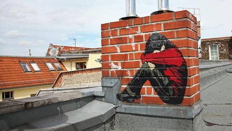 Ein Bild des Street-Art-Künstlers "Alias" auf einem Schornstein auf einem Dach - ein Junge, der sein Gesicht in seine Arme presst.