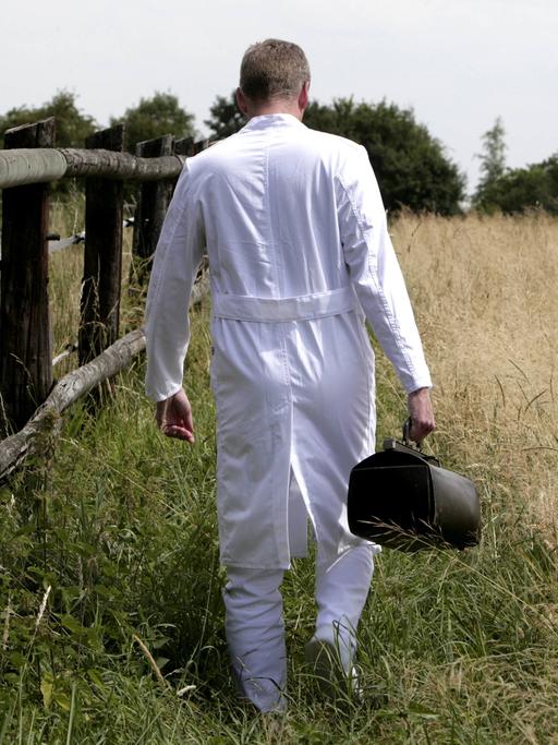 Ein Landarzt im weißen Kittel mit Ledertasche geht zwischen einem Pferdegatter und einem Kornfeld.
