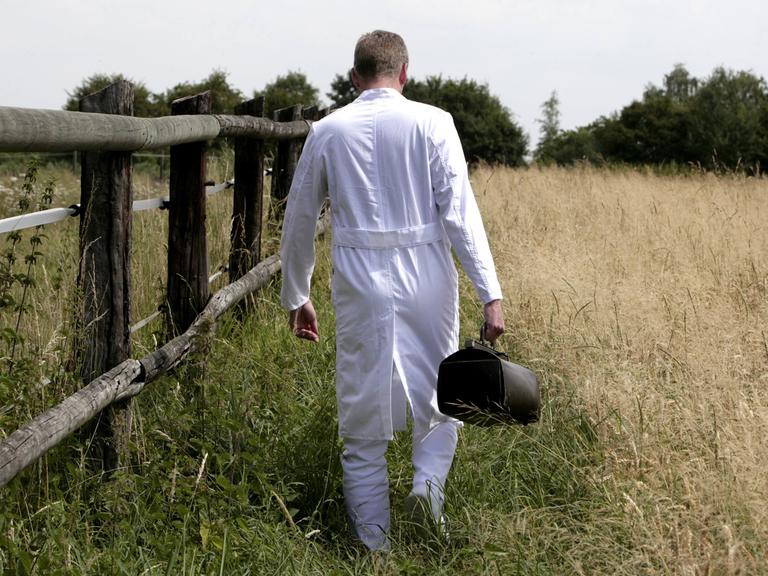 Ein Landarzt im weißen Kittel mit Ledertasche geht zwischen einem Pferdegatter und einem Kornfeld.