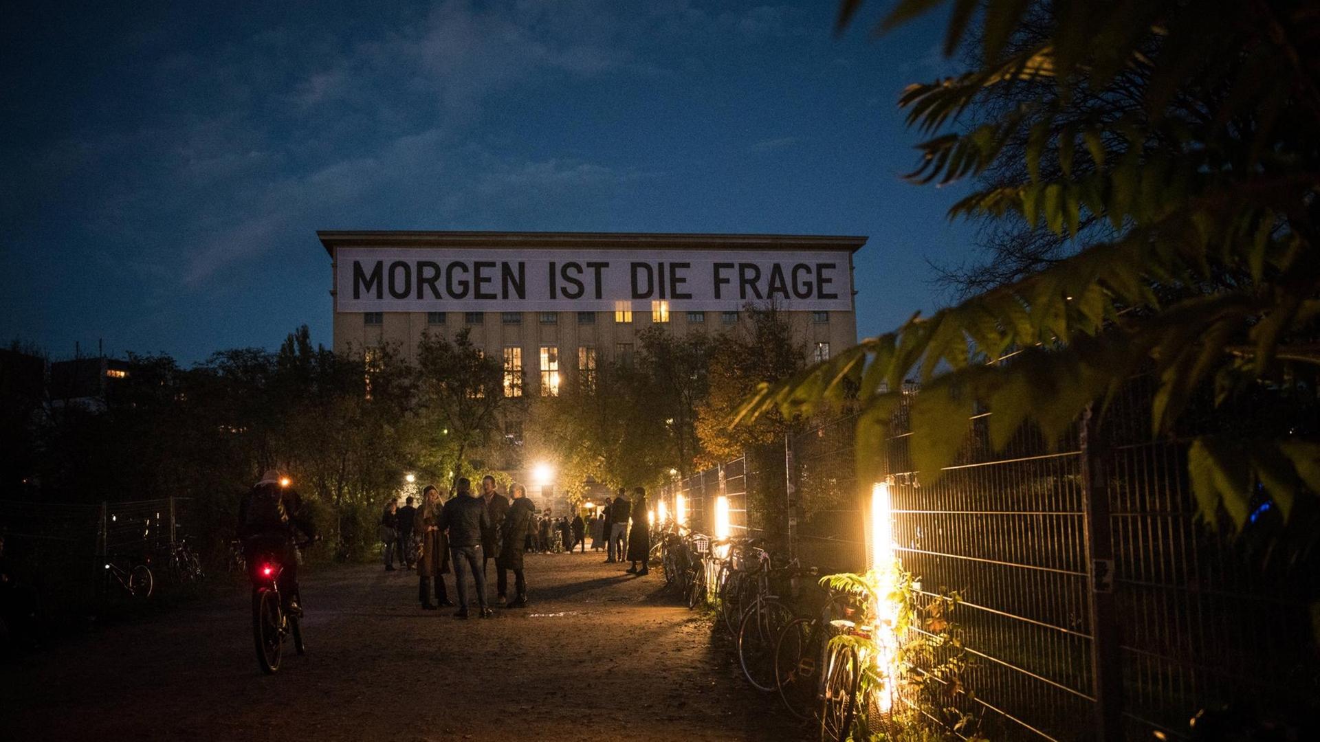 Der Schriftzug "Morgen ist die Frage" des Künstlers Rirkrit Tiravanija hängt am 1. November 2020 zu Beginn des neuerlichen Lockdowns an der Fassade des Techno Clubs Berghain in Berlin-Friedrichshain. Im Dämmerlicht stehen Menschen vor dem Gebäude. Der Schriftzug "Morgen ist die Frage" des Künstlers Rirkrit Tiravanija hängt am 1. November 2020 zu Beginn des neuerlichen Lockdowns an der Fassade des Techno Clubs Berghain in Berlin-Friedrichshain. Im Dämmerlicht stehen Menschen vor dem Gebäude.