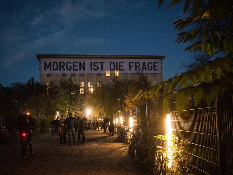 Der Schriftzug "Morgen ist die Frage" des Künstlers Rirkrit Tiravanija hängt am 1. November 2020 zu Beginn des neuerlichen Lockdowns an der Fassade des Techno Clubs Berghain in Berlin-Friedrichshain. Im Dämmerlicht stehen Menschen vor dem Gebäude.