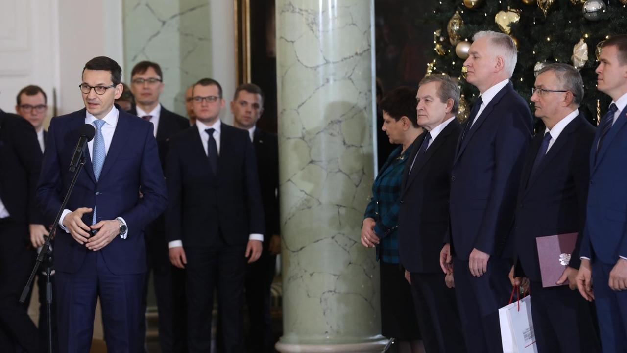 Der polnische Ministerpräsident Mateusz Morawiecki (l) setht am 9. Januar 2018 in einem Marmorsaal. Neben ihm stehen mehrere Männer in dunklen Anzügen.|