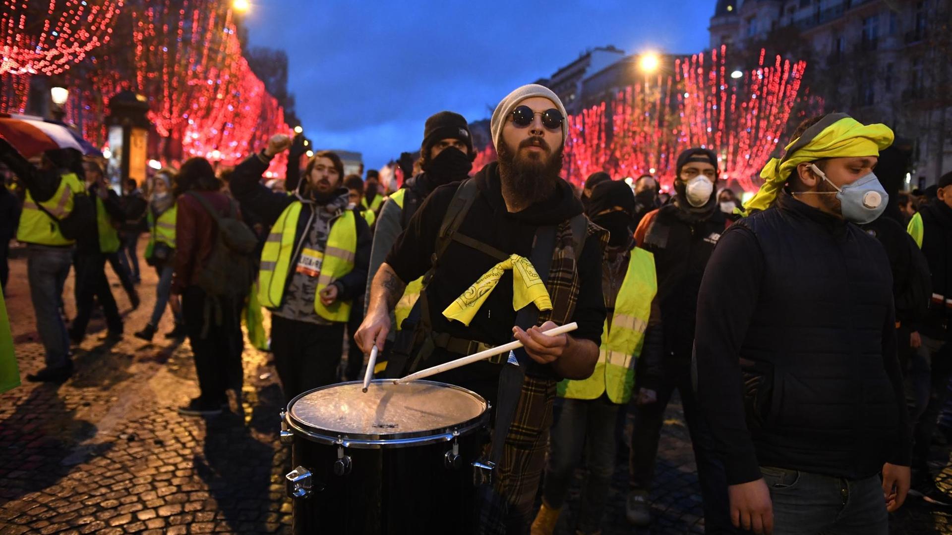 Durch eine mit roten Lichtern gerahmte Straße laufen einige Gelbwesten. Der Mann im Vordergrund trägt eine Trommel. Durch eine mit roten Lichtern gerahmte Straße laufen einige Gelbwesten. Der Mann im Vordergrund trägt eine Trommel.