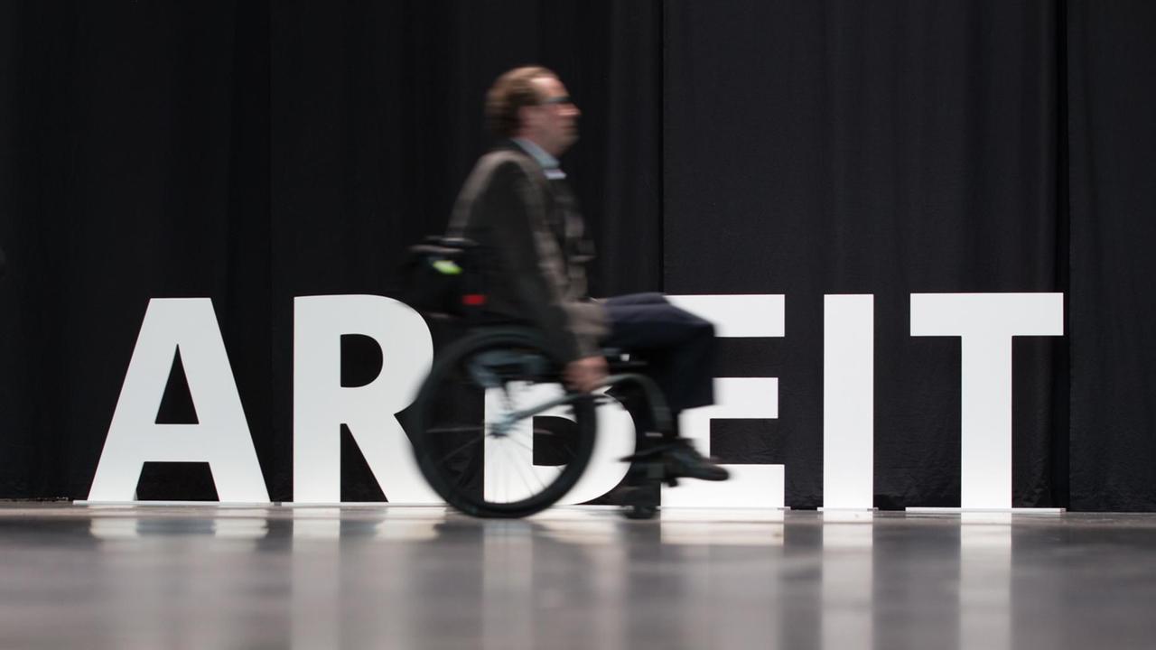 Ein Mensch mit Behinderung fährt am 20.02.2017 in Bielefeld mit seinem Rollstuhl an dem aufgestellten Wort "Arbeit" entlang. Ein Mensch mit Behinderung fährt am 20.02.2017 in Bielefeld mit seinem Rollstuhl an dem aufgestellten Wort "Arbeit" entlang.