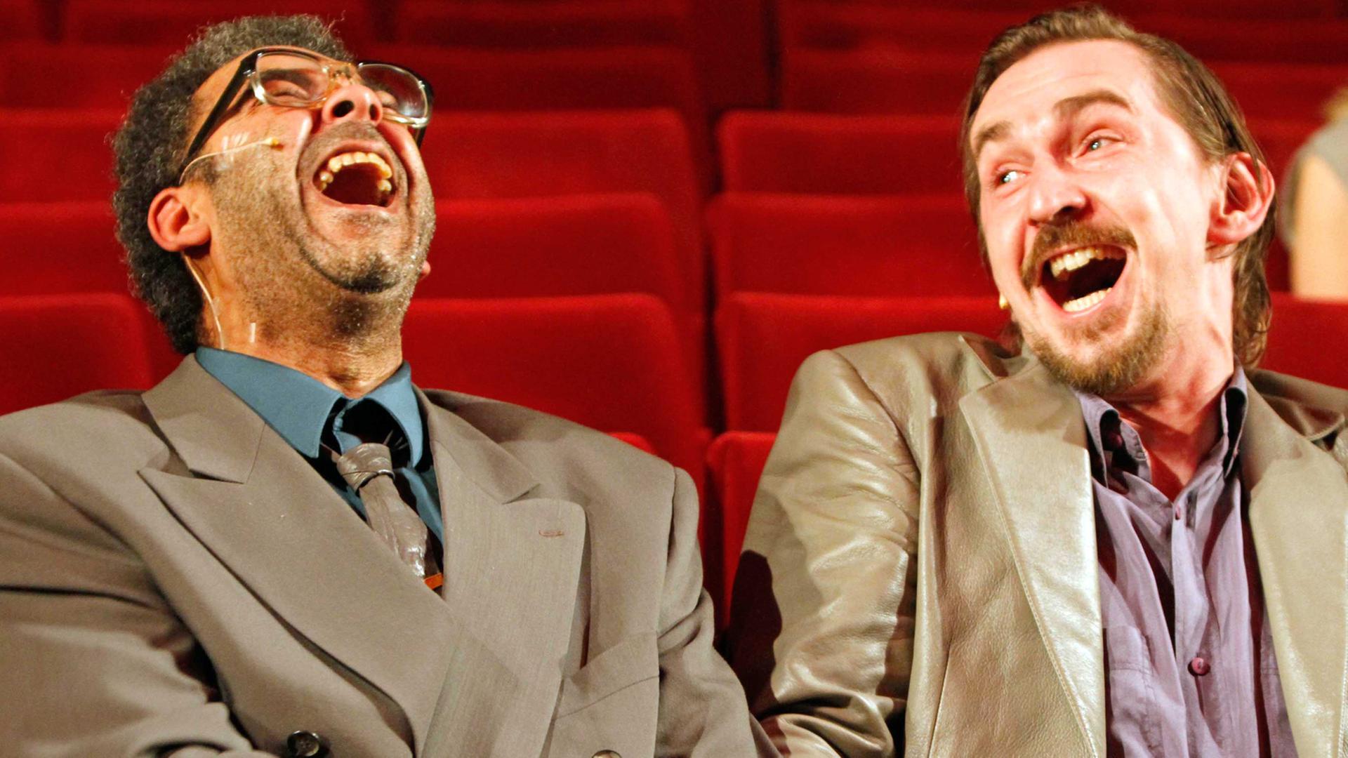 Die Schauspieler Falilou Seck (links) und Till Wonka sitzen in einer Kinositzreihe mit roten Stühlen nebeneinander, geben sich die Hand und werfen beim Lachen den Kopf in den Nacken.