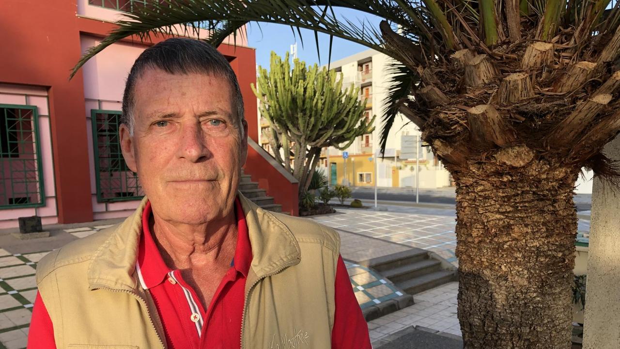 Ein älterer Herr mit rotem T-Shirt und Jacke steht vor einer großen Palme in der Sonne.