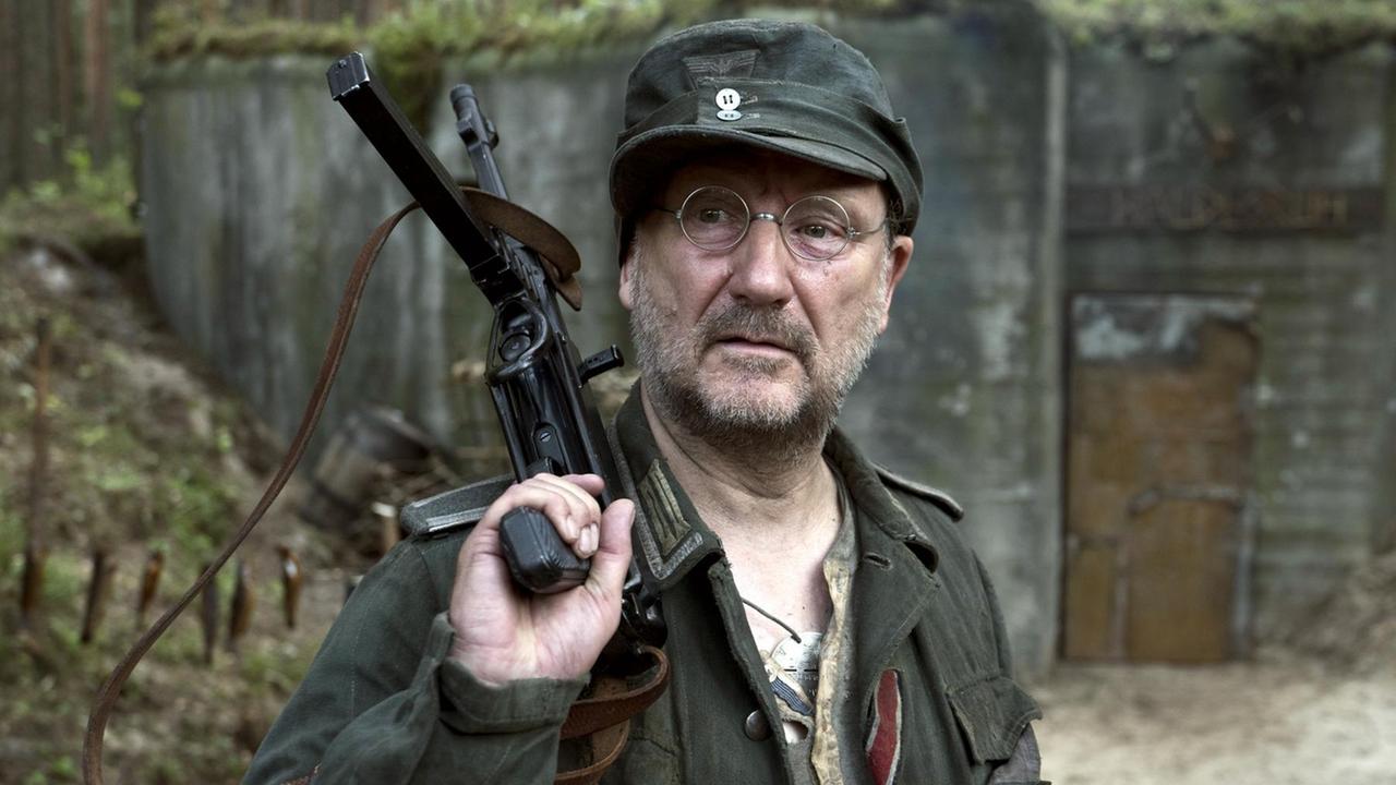 Rainer Bock in der Rolle als Soldat, der lässig eine Waffe schultert.