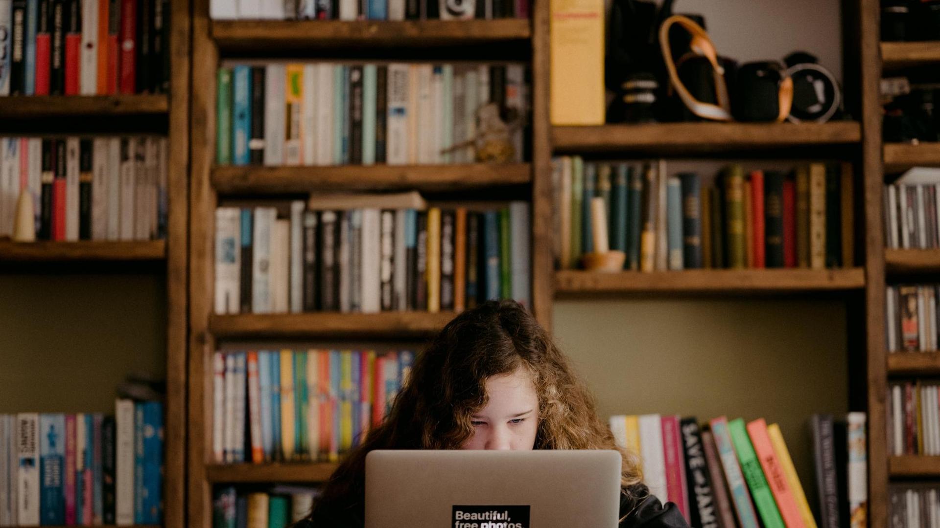 Ein Mädchen sitzt vor einer Bücherwand vor seinem Laptop.